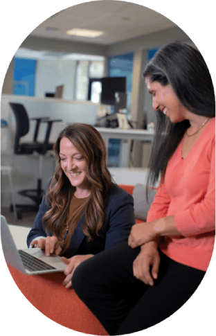 Office discussion – Two women laughing while using laptop.